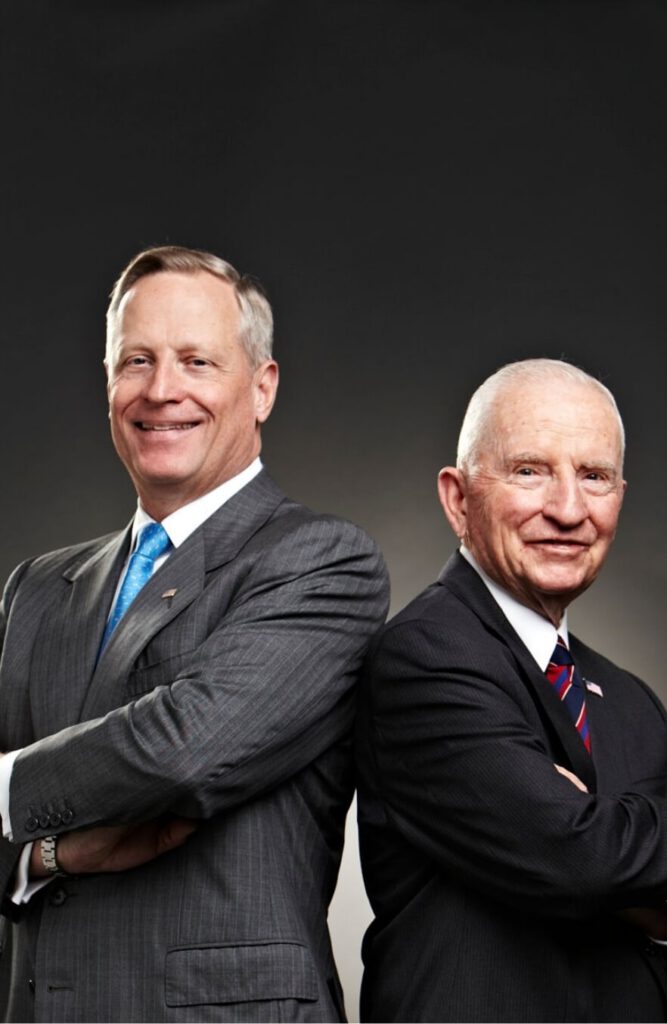 Ross Perot Jr. and Ross Perot Sr. standing back-to-back and smiling.