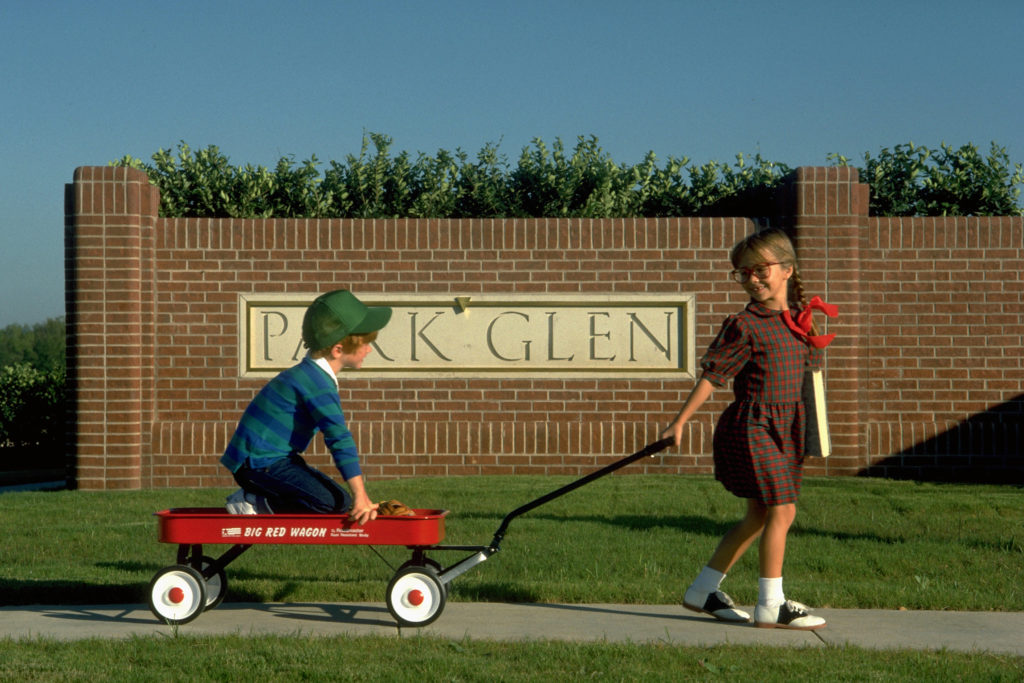 A young girl toting along her brother in a wagon in front of the Park Glen entrance sign